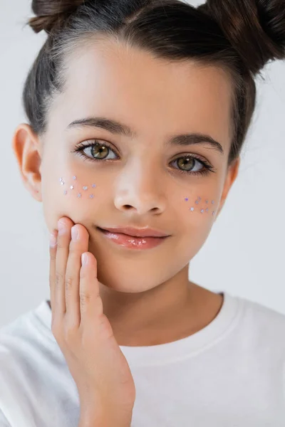 Портрет усміхненої дівчини з блиском губ та блискучими зірками на обличчі, що торкаються щоки, ізольовані на сірому — стокове фото
