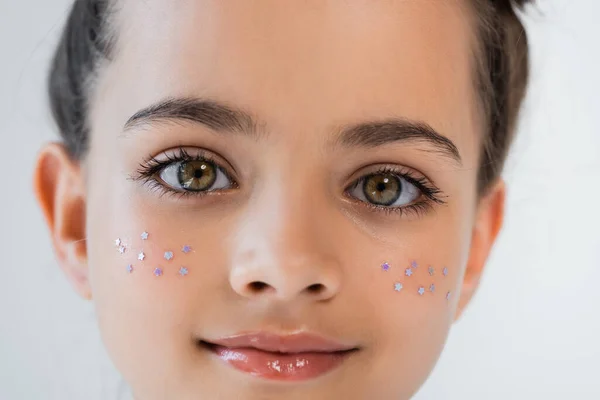 Portrait of smiling girl with hazel eyes and sparkling glitter stars on cheeks looking at camera isolated on grey — Fotografia de Stock