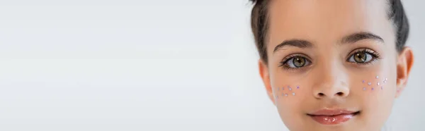 Portrait of smiling girl with hazel eyes and sparkling glitter stars on face looking at camera isolated on grey, banner — Stock Photo