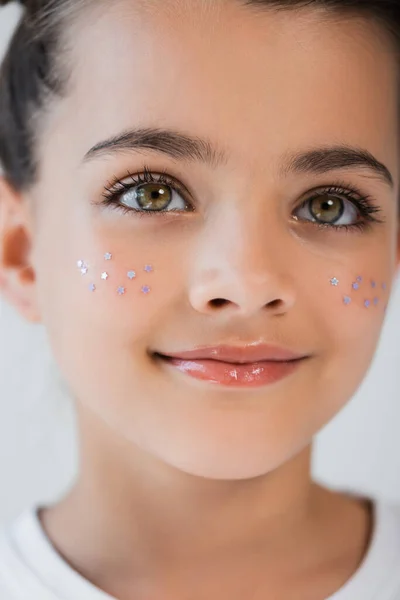 Close up view of cheerful girl with hazel eyes and sparkling glitter stars on cheeks isolated on grey — Foto stock
