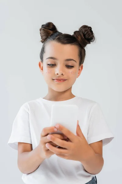 Cheerful girl with glitter stars on cheeks using smartphone isolated on grey — стоковое фото