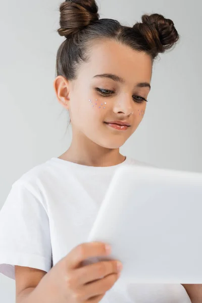 Cheerful girl with glitter stars on cheeks smiling while using digital tablet isolated on grey — Photo de stock