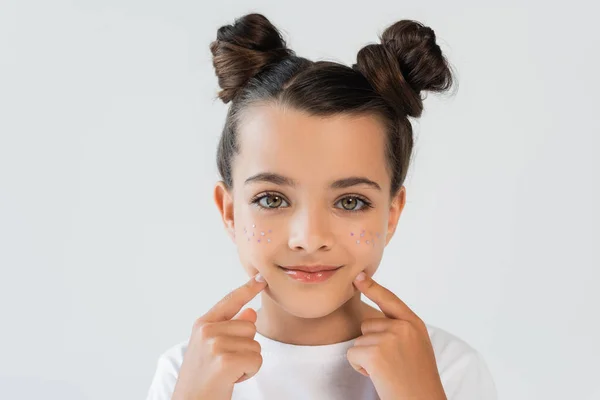 Portrait of cheerful girl with lip gloss and sparkling glitter stars pointing at face isolated on grey - foto de stock