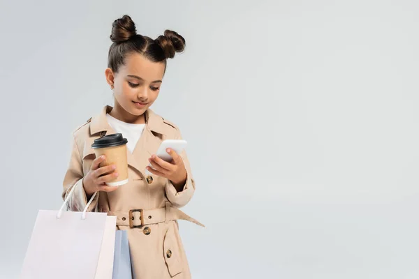 Cute girl in trench coat holding paper cup and shopping bags while using smartphone isolated on grey — Stock Photo