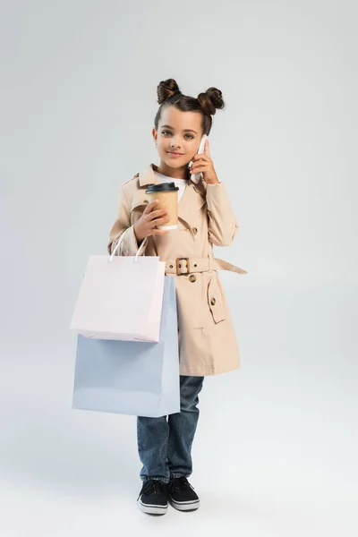 Щаслива дівчина в траншеї пальто і джинси тримає паперовий стаканчик і сумки під час розмови на смартфоні на сірому — стокове фото