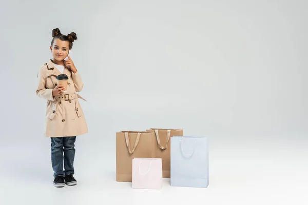 Happy girl in trench coat and jeans holding paper cup and talking on smartphone near shopping bags on grey — Stockfoto