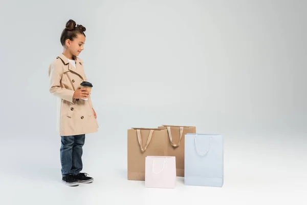 Happy girl in trench coat and jeans holding paper cup and looking at shopping bags on grey — Foto stock