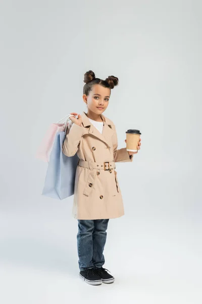 Cute girl in trench coat and jeans holding takeaway drink and shopping bags on grey — Stock Photo