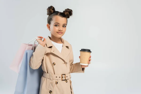 Adorable girl in trench coat holding takeaway drink and shopping bags isolated on grey - foto de stock