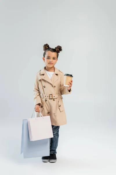Мила дівчина в стильному тренч пальто і джинси тримає винос напій і сумки для покупок на сірому — стокове фото