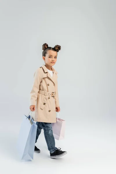 Full length of stylish kid in trench coat and jeans holding shopping bags and walking on grey, black friday concept — Stockfoto