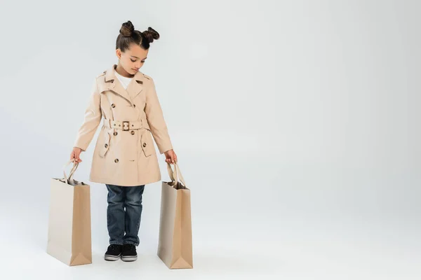 Full length of stylish kid in trench coat and jeans holding shopping bags on grey, black friday concept - foto de stock