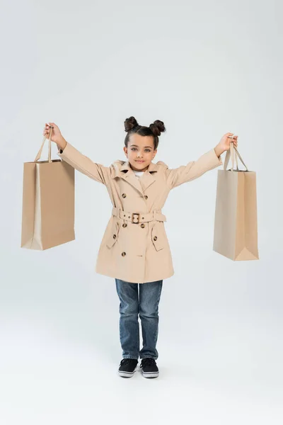 Full length of cheerful kid in trench coat and jeans holding shopping bags on grey, black friday concept — Foto stock