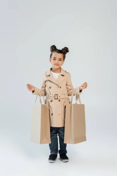 Full length of stylish child in trench coat and jeans holding shopping bags on grey, black friday concept — стоковое фото