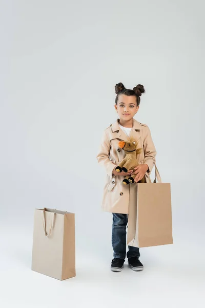 Full length of cheerful kid in trench coat and jeans holding soft toy and shopping bag on grey — Stockfoto