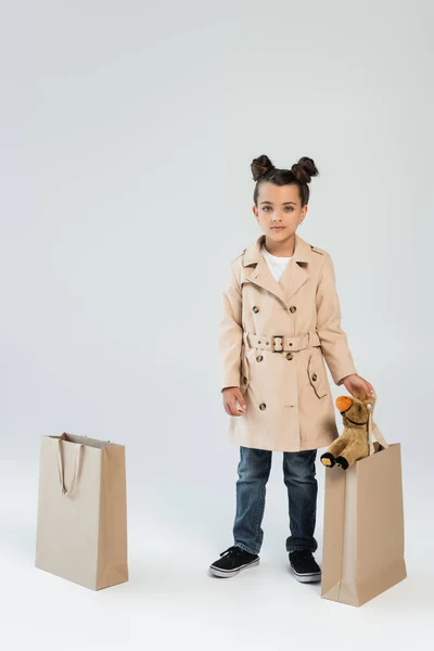 Повна довжина стильної дитини в траншеї пальто і джинси, що тримає сумку з іграшковим конем на сірому — стокове фото