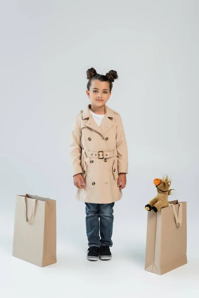 Full length of cheerful kid in trench coat and jeans standing near shopping bags with soft toy on grey — Fotografia de Stock