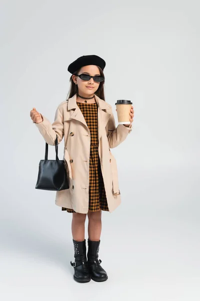 Full length of adorable girl in trench coat and sunglasses holding takeaway drink and handbag on grey — Photo de stock