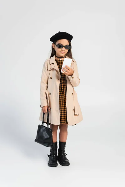 Full length of adorable girl in trench coat and beret holding smartphone and handbag on grey - foto de stock