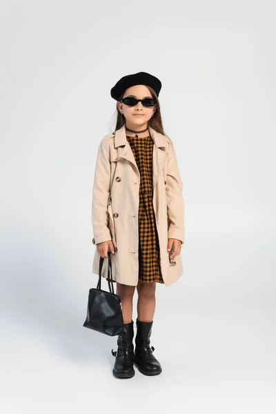 Full length of stylish girl in trench coat and beret holding handbag on grey — Stockfoto