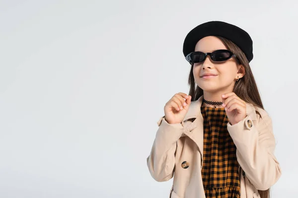 Happy girl in stylish trench coat and sunglasses smiling isolated on grey — Stockfoto