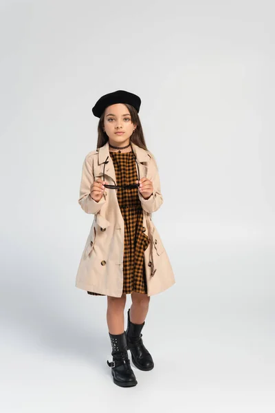 Full length of cute girl in stylish trench coat and beret holding sunglasses on grey - foto de stock