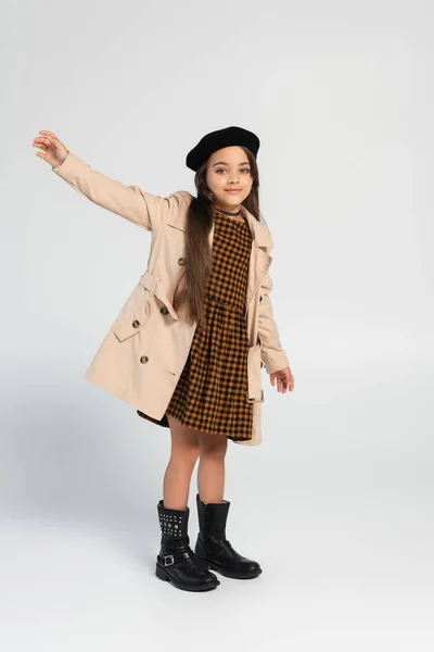 Full length of cheerful girl in stylish trench coat and beret gesturing while standing on grey — Stock Photo