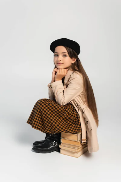 Full length of cute kid in stylish autumnal outfit and beret sitting on stacked books on grey — стоковое фото