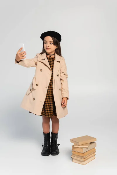 Full length of cute kid in stylish autumnal outfit and beret taking selfie near stacked books on grey - foto de stock