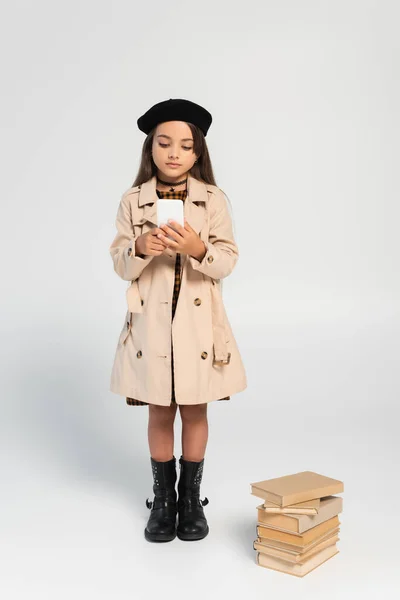 Full length of cute kid in stylish autumnal outfit and beret using smartphone near stacked books on grey — Stock Photo