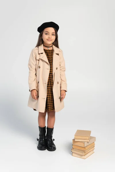 Full length of smiling kid in stylish autumnal outfit and beret standing near stacked books on grey — Stockfoto