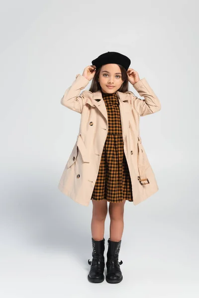 Full length of cheerful girl in stylish trench coat adjusting beret while standing on grey — Stockfoto