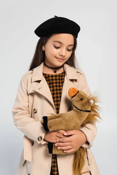 Cheerful girl in stylish trench coat and beret standing with toy horse isolated on grey — стоковое фото