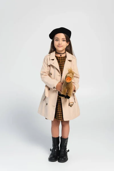 Full length of cute girl in stylish trench coat and beret standing with toy horse on grey — стоковое фото