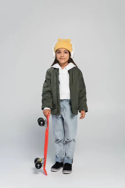 Full length of cheerful girl in headphones and trendy autumnal outfit holding penny board on grey — Stockfoto
