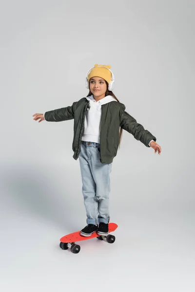 Full length of cheerful girl in wireless headphones and trendy autumnal outfit riding penny board on grey — Stock Photo