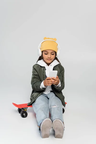 Full length of cute girl in wireless headphones and trendy outfit using smartphone and sitting on penny board on grey — Stock Photo