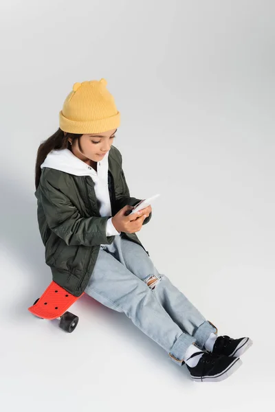 High angle view of cute girl in trendy autumnal outfit using smartphone and sitting on penny board on grey — Photo de stock