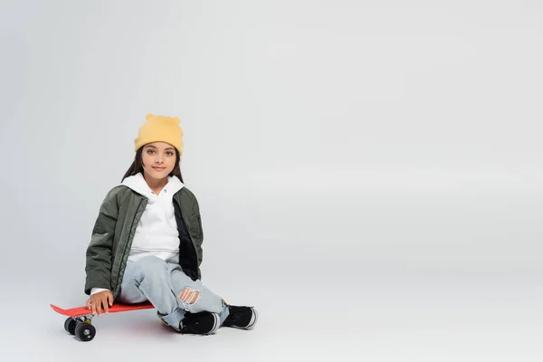 Full length of cute kid in trendy autumnal outfit smiling while sitting on penny board on grey — Fotografia de Stock