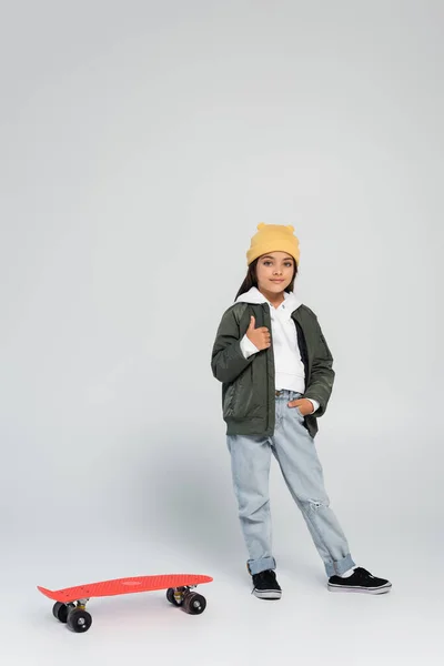 Full length of cute girl in trendy outfit standing with hand in pocket and showing thumb up near penny board on grey — Stockfoto