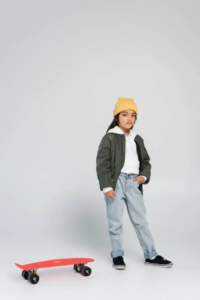 Full length of cute girl in trendy autumnal outfit standing with hand in pocket near penny board on grey — Stock Photo