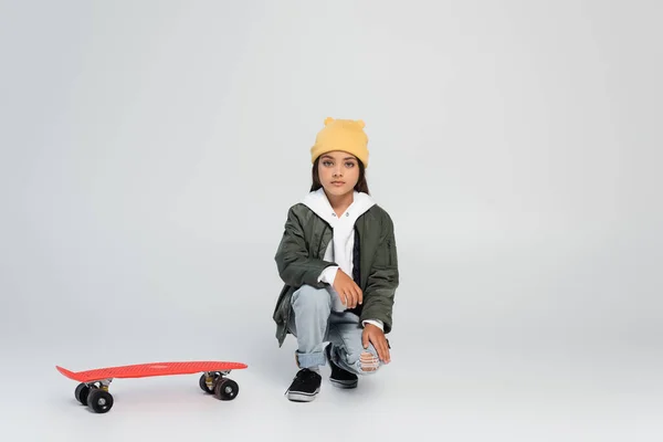 Full length of cute girl in trendy autumnal outfit sitting near penny board on grey - foto de stock