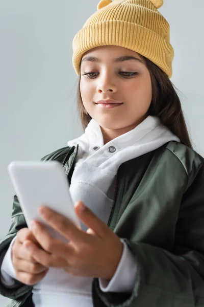 Cheerful kid in trendy autumnal outfit and beanie hat using smartphone isolated on grey — Stock Photo