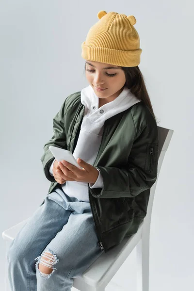 High angle view of cheerful kid in trendy autumnal outfit sitting on high chair and using smartphone isolated on grey — Stockfoto