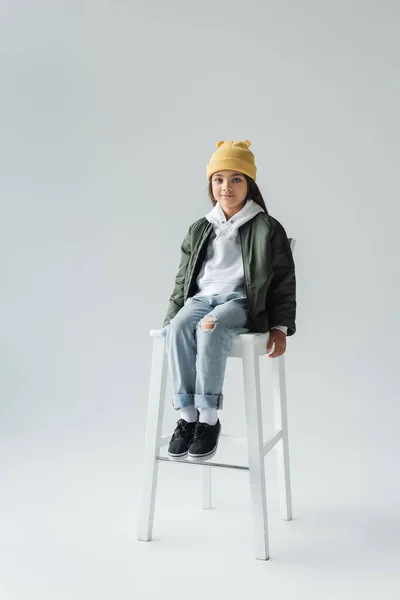 Full length of cute girl in trendy autumnal outfit sitting on high chair on grey - foto de stock