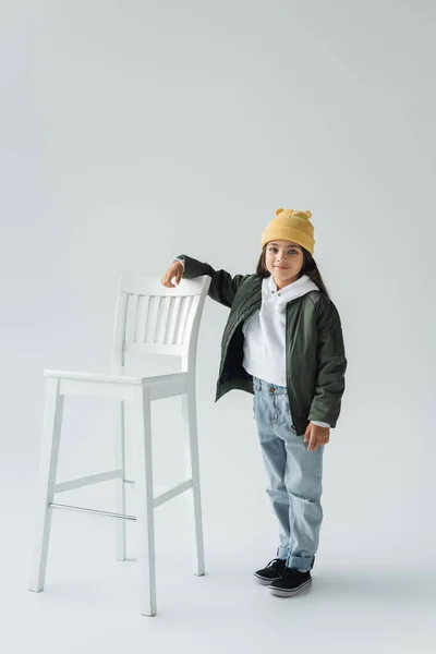 Full length of cheerful girl in trendy autumnal outfit and beanie hat standing near high chair on grey — Foto stock
