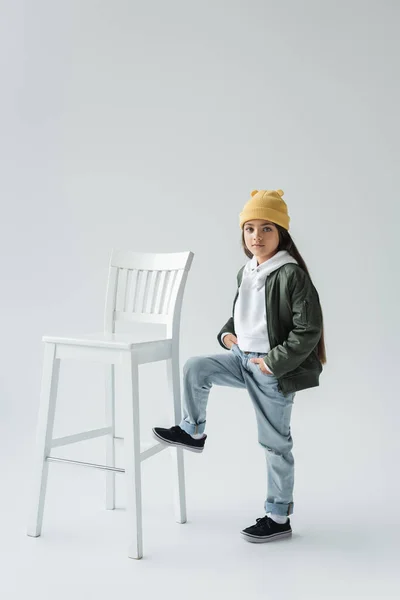 Full length of cute girl in trendy autumnal outfit and beanie hat standing with hands in pockets near high chair on grey - foto de stock