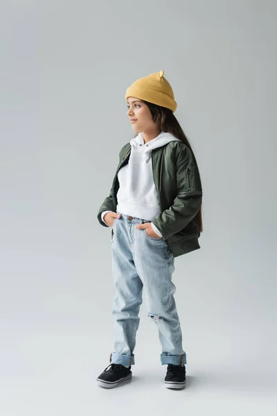 Full length of smiling kid in yellow beanie hat and stylish autumnal outfit posing with hands in pockets on grey - foto de stock