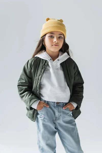 Portrait of cute kid in yellow beanie hat and stylish autumnal outfit posing with hands in pockets isolated on grey — Foto stock