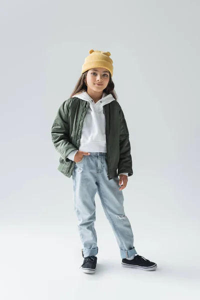 Full length of cute girl in beanie hat and stylish autumnal outfit posing with hand in pocket on grey — стоковое фото